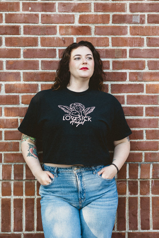 A person wearing a black crop top with an angel graphic and the word 'lovesick angel' on it, paired with blue jeans, standing against a brick wall background.