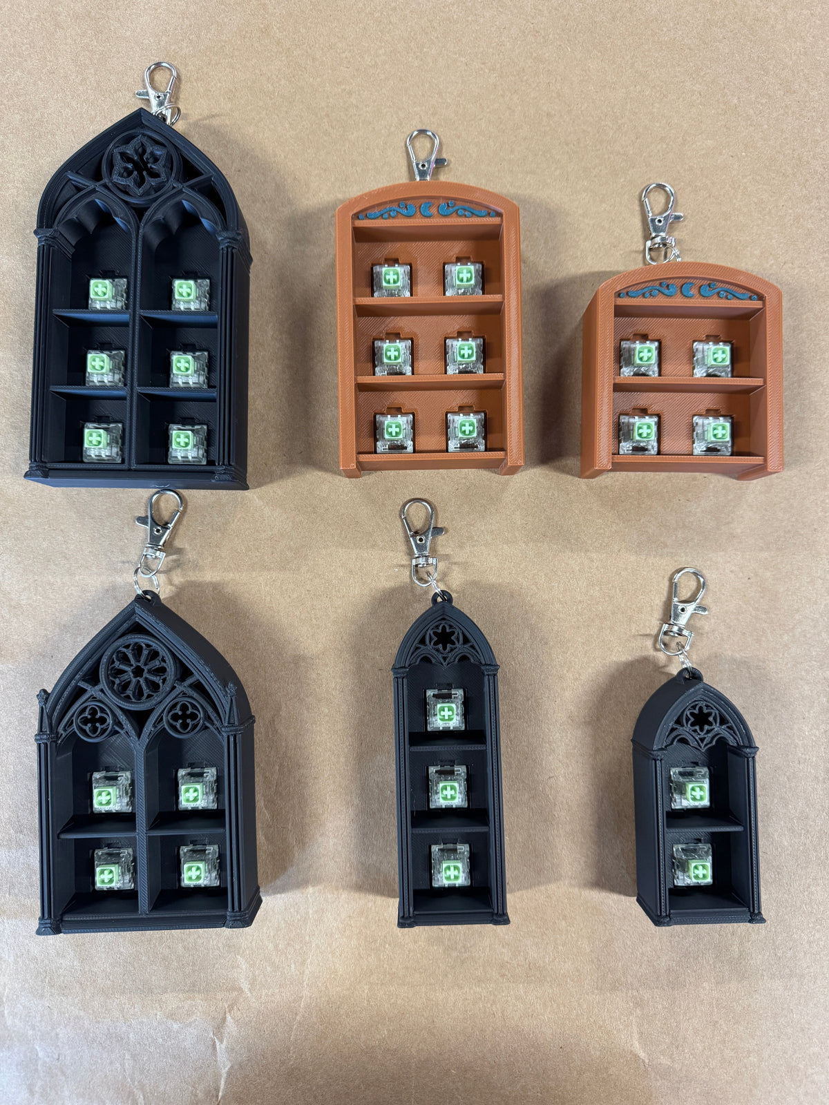 Six small 3D-printed keychain organizers in two colors: two terracotta orange and four black, each with a gothic-style window design, internal compartments, and a silver lobster clasp on a tan background.