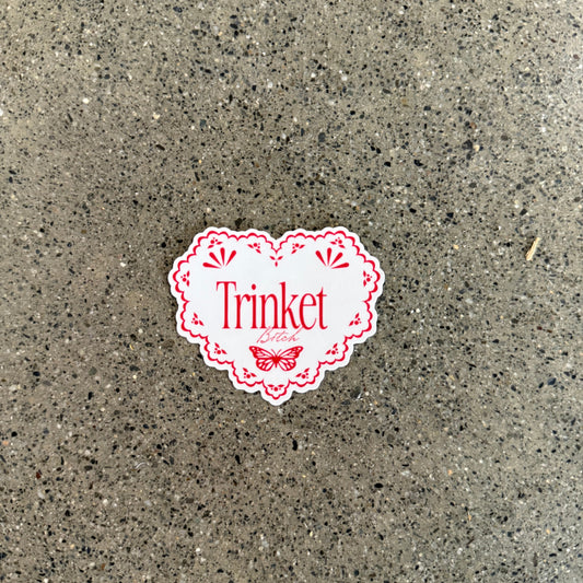 Sticker of a scallop edged heart that reads "Trinket Bitch" in a serif and script font with a butterfly. Text is in red on a white background